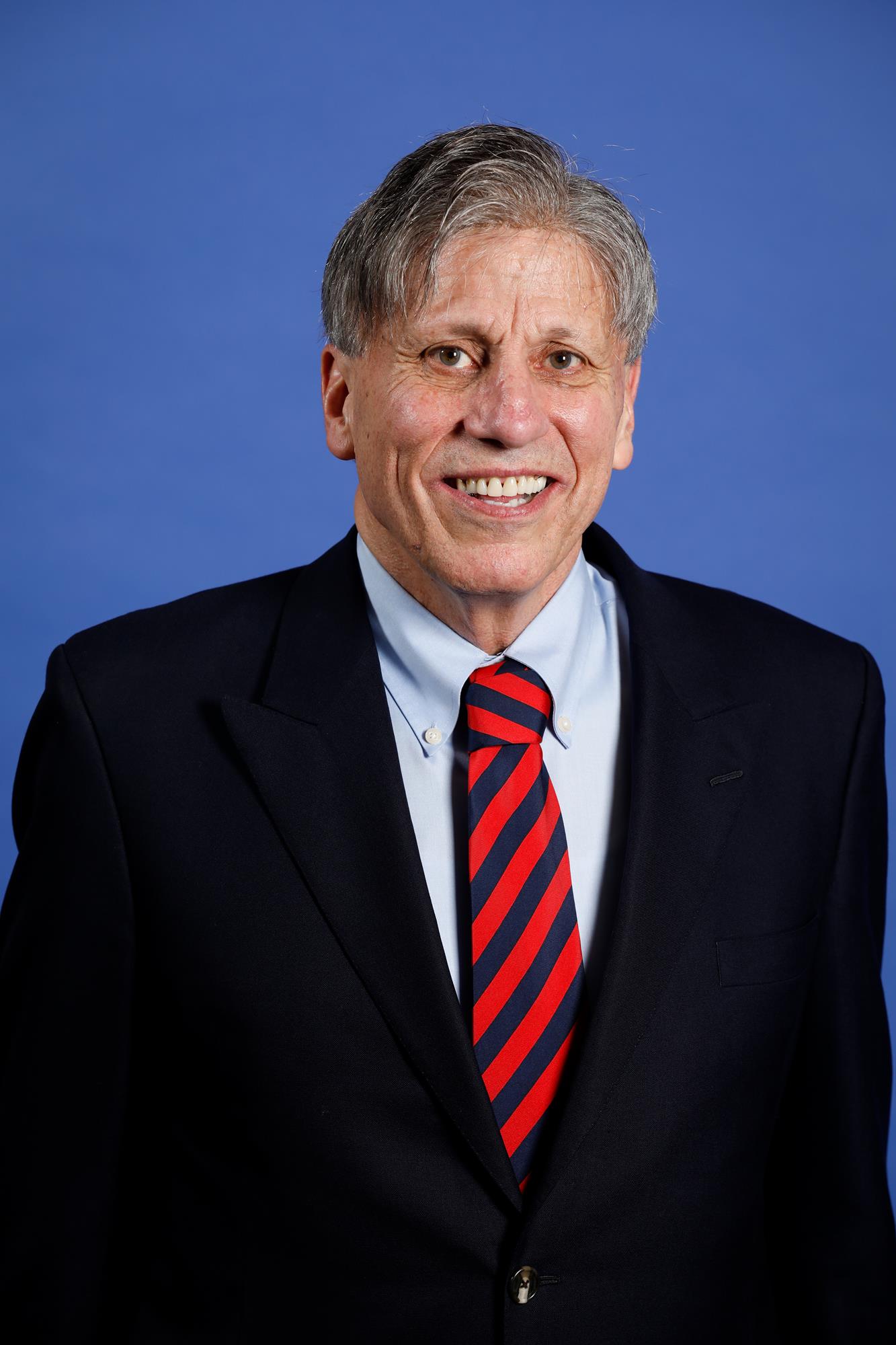 A man in a suit and tie smiling at a blue background.